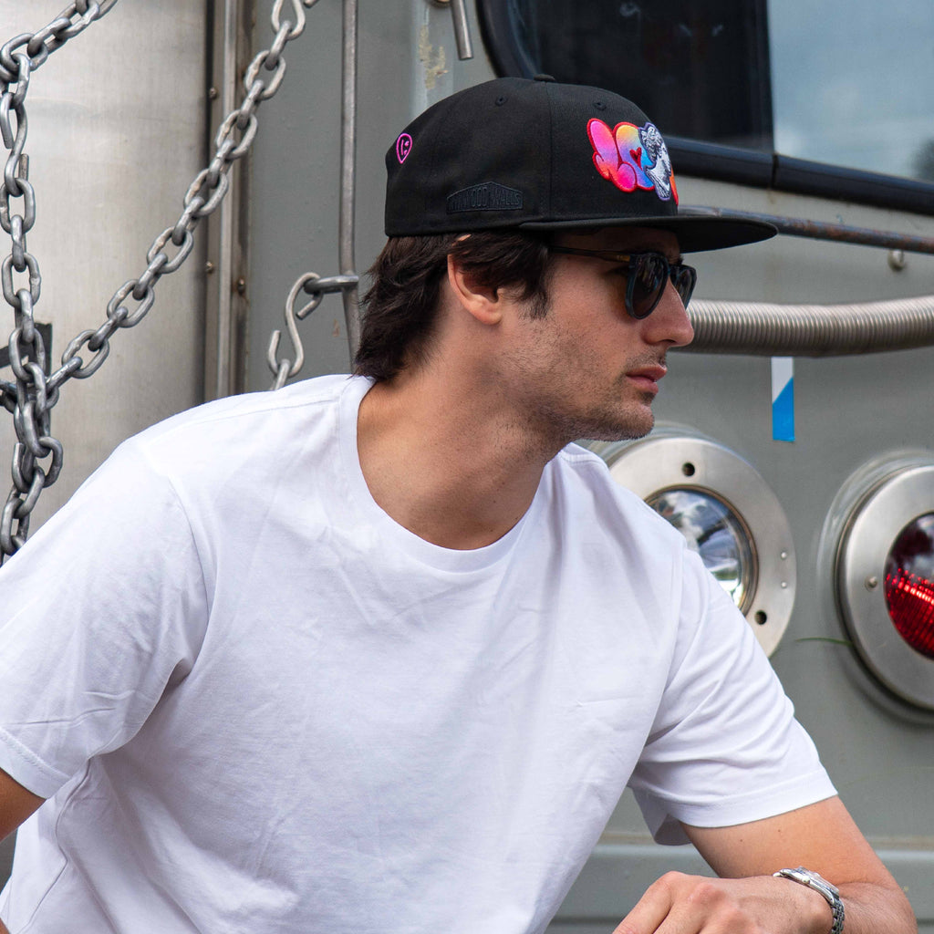 Man wearing a black cap with colorful text and sunglasses, standing in front of a large truck.