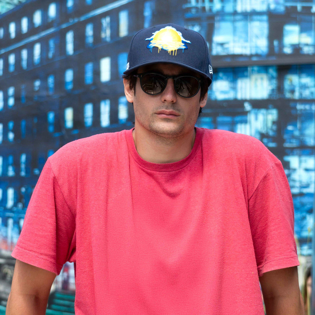 Man wearing a red shirt, dark cap, and sunglasses with a cityscape background