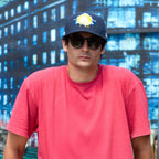 Man wearing a red shirt, dark cap, and sunglasses with a cityscape background