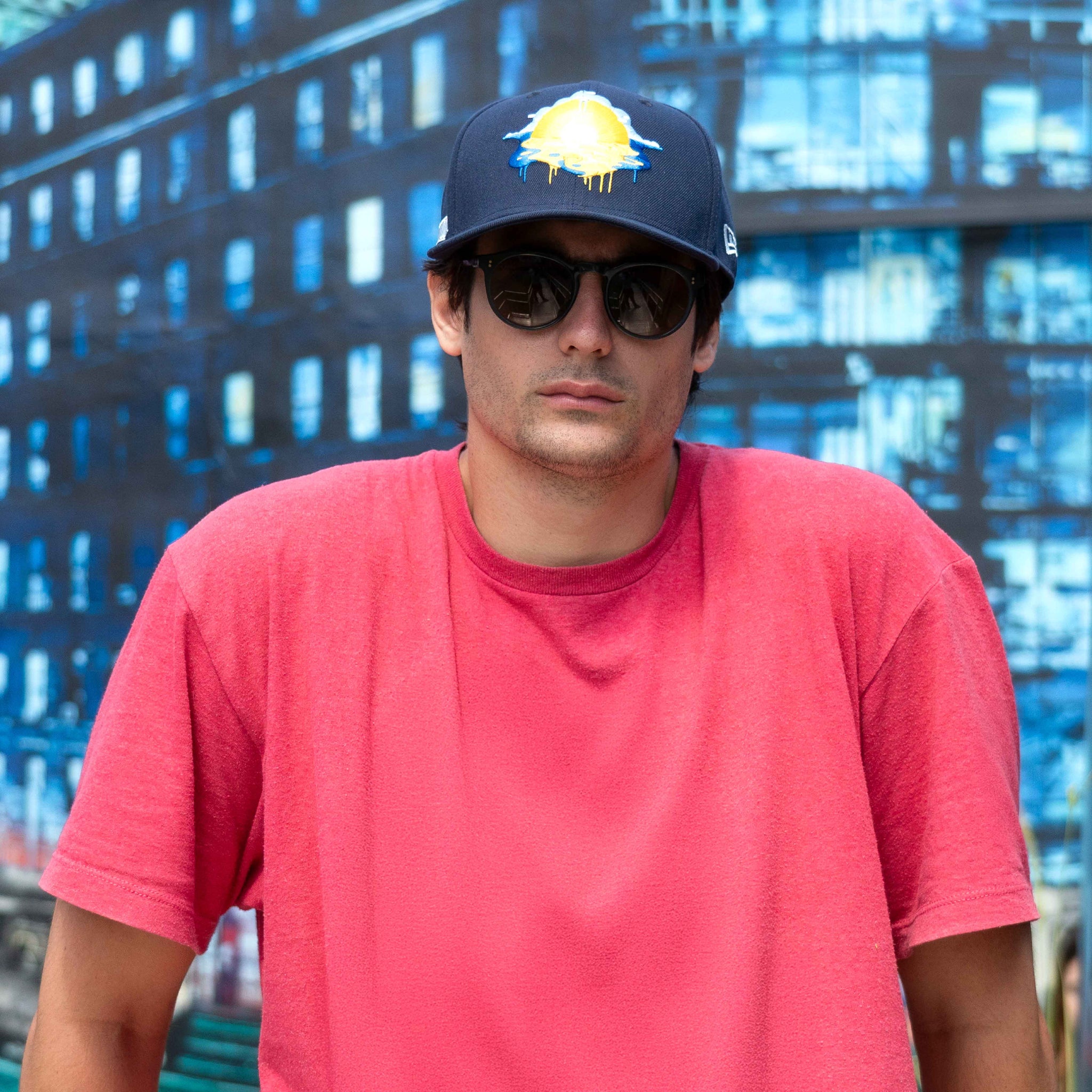 Man wearing a red shirt, dark cap, and sunglasses with a cityscape background
