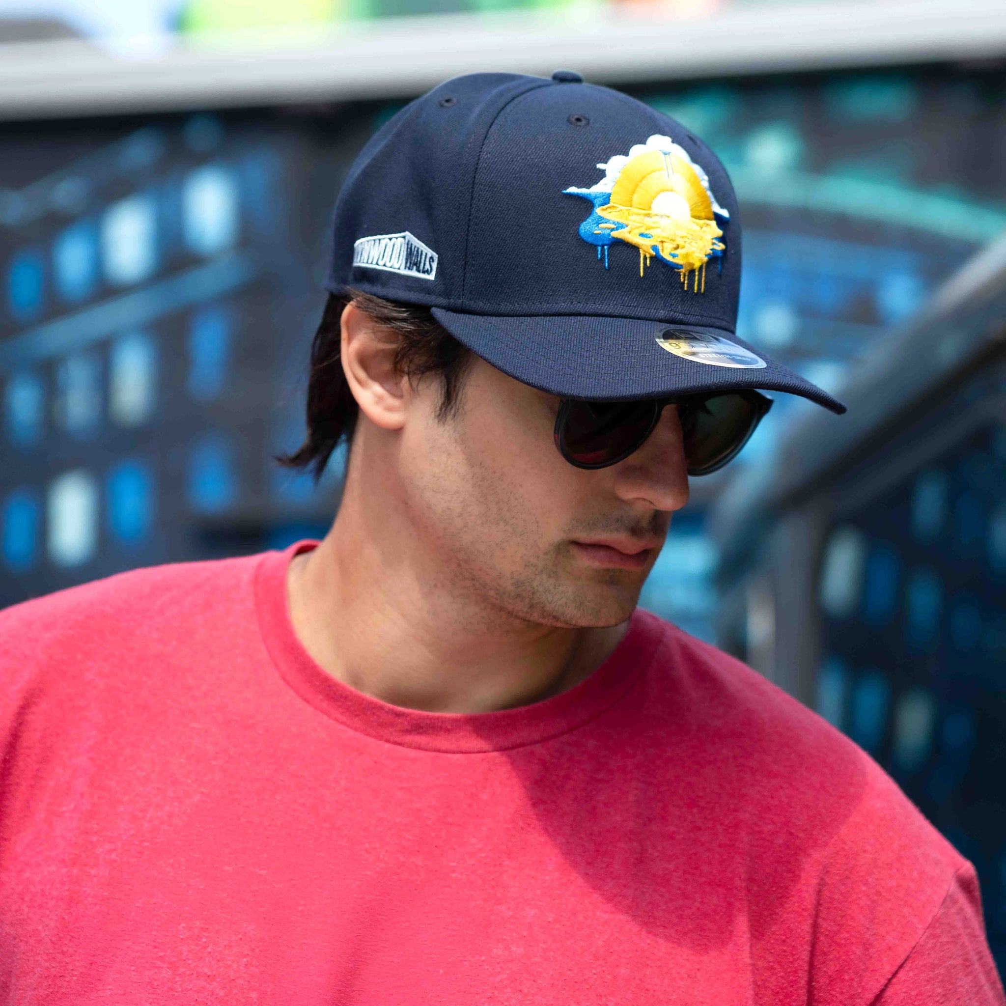 Man wearing a navy cap with a logo and sunglasses, standing in front of a blurred background