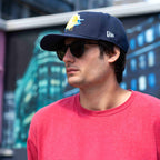 Man wearing a navy cap with a logo and sunglasses, standing in front of a blurred background