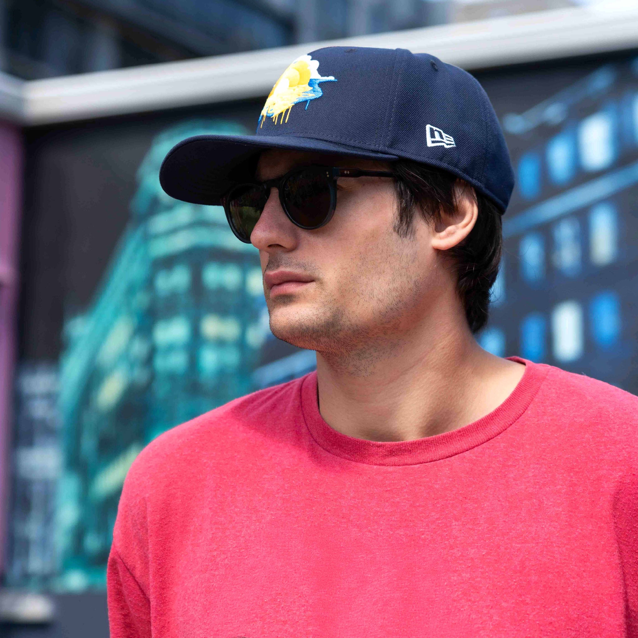 Man wearing a navy cap with a logo and sunglasses, standing in front of a blurred background